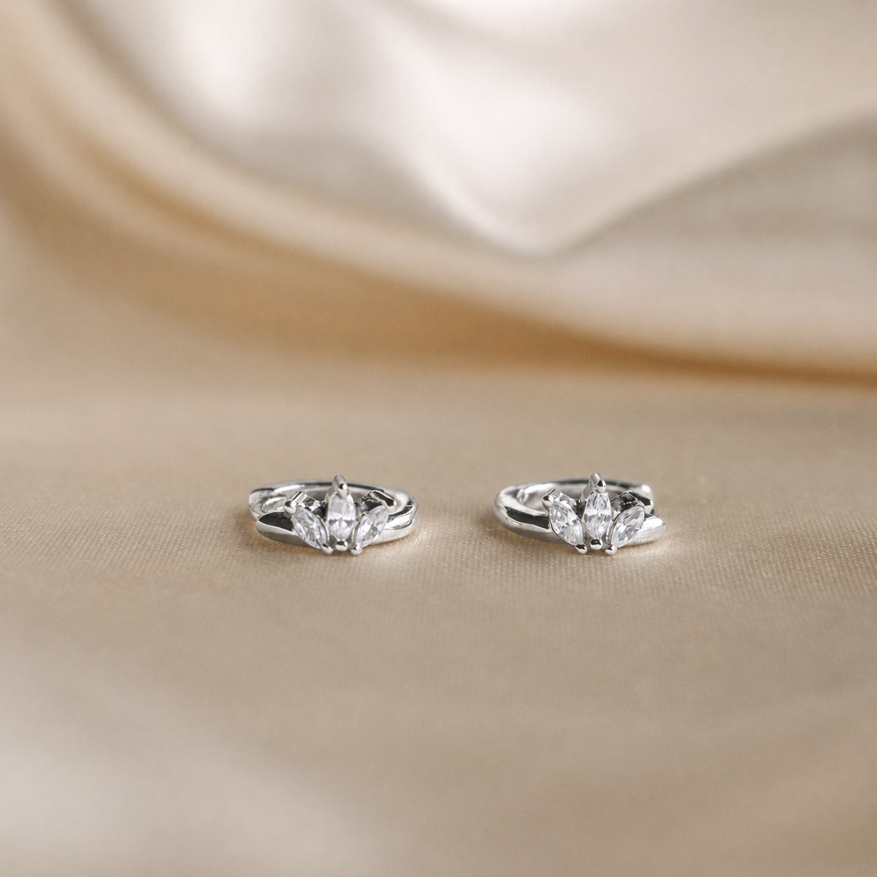 A pair of silver-toned bridal rings with marquise-cut center stones and smaller side stones, displayed on a soft beige satin fabric background.